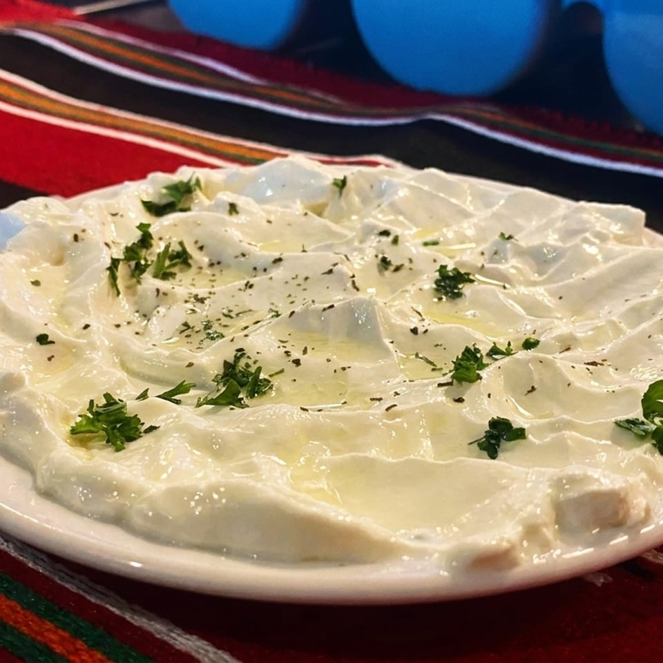 A white plate of labneh, drizzled with olive oil and garnished with herbs, on a red patterned tablecloth.